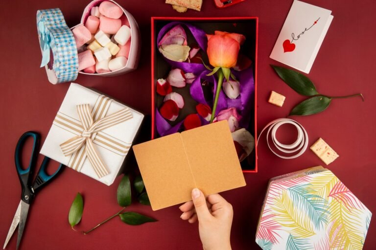 top-view-female-hand-with-small-open-postcard-gift-box-with-coral-color-rose-flower-with-scattered-petals-box-filled-with-marshmallow-red-table_141793-8844